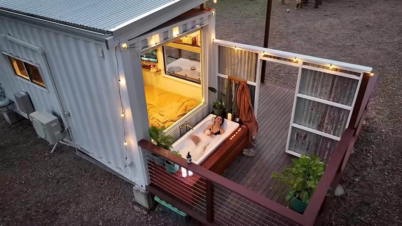 Outdoor bathtub built into the timber deck of a white shipping container home at dusk, with candles and string lights