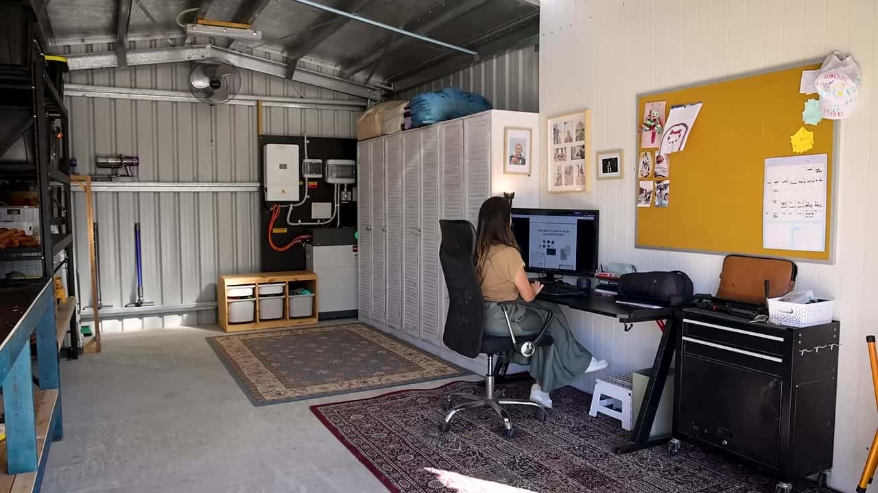 Interior of a metal shed with an office desk, white wardrobes, storage shelves, and off-grid electrical gear on the wall