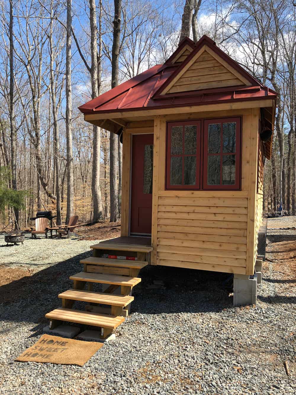 Front of Ryan Mitchell's tiny house with wooden entry steps, red-trimmed double windows, and a steep red metal roof in a wooded lot