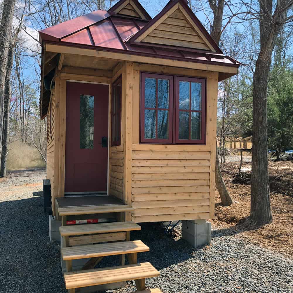 Ryan Mitchell's cedar-sided tiny house on wheels with a red standing-seam metal roof, cross gables, and a dark red front door