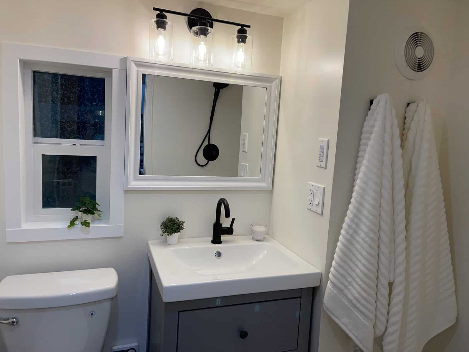 Bathroom with a gray vanity and white sink, black faucet, framed mirror, three-light fixture, a window, and a toilet