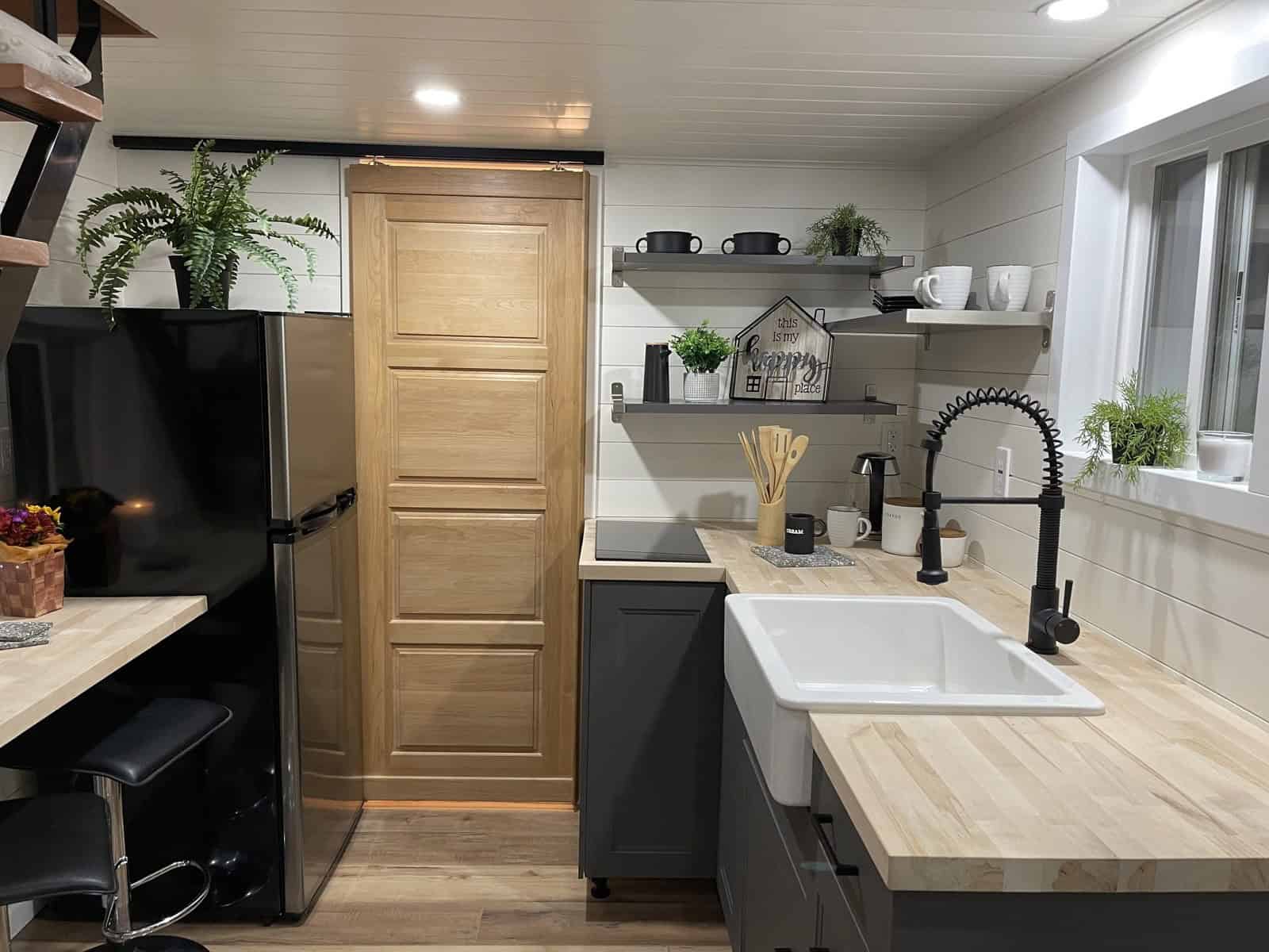 Kitchen with a white farm sink and black faucet, butcher block counters, gray lower cabinets, a two-burner cooktop, open shelves, a stainless refrigerator, and a wood barn door