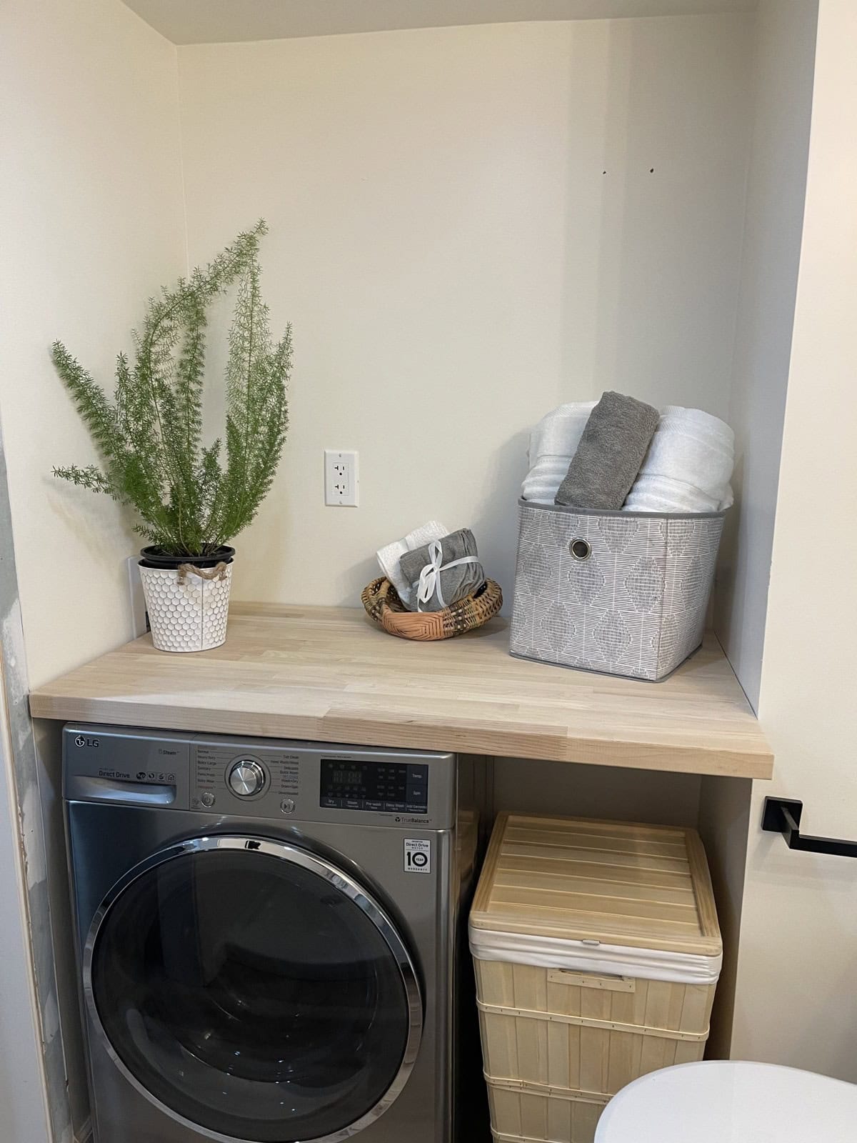 Front-load washer under a butcher block shelf with baskets and a plant, beside a wood hamper