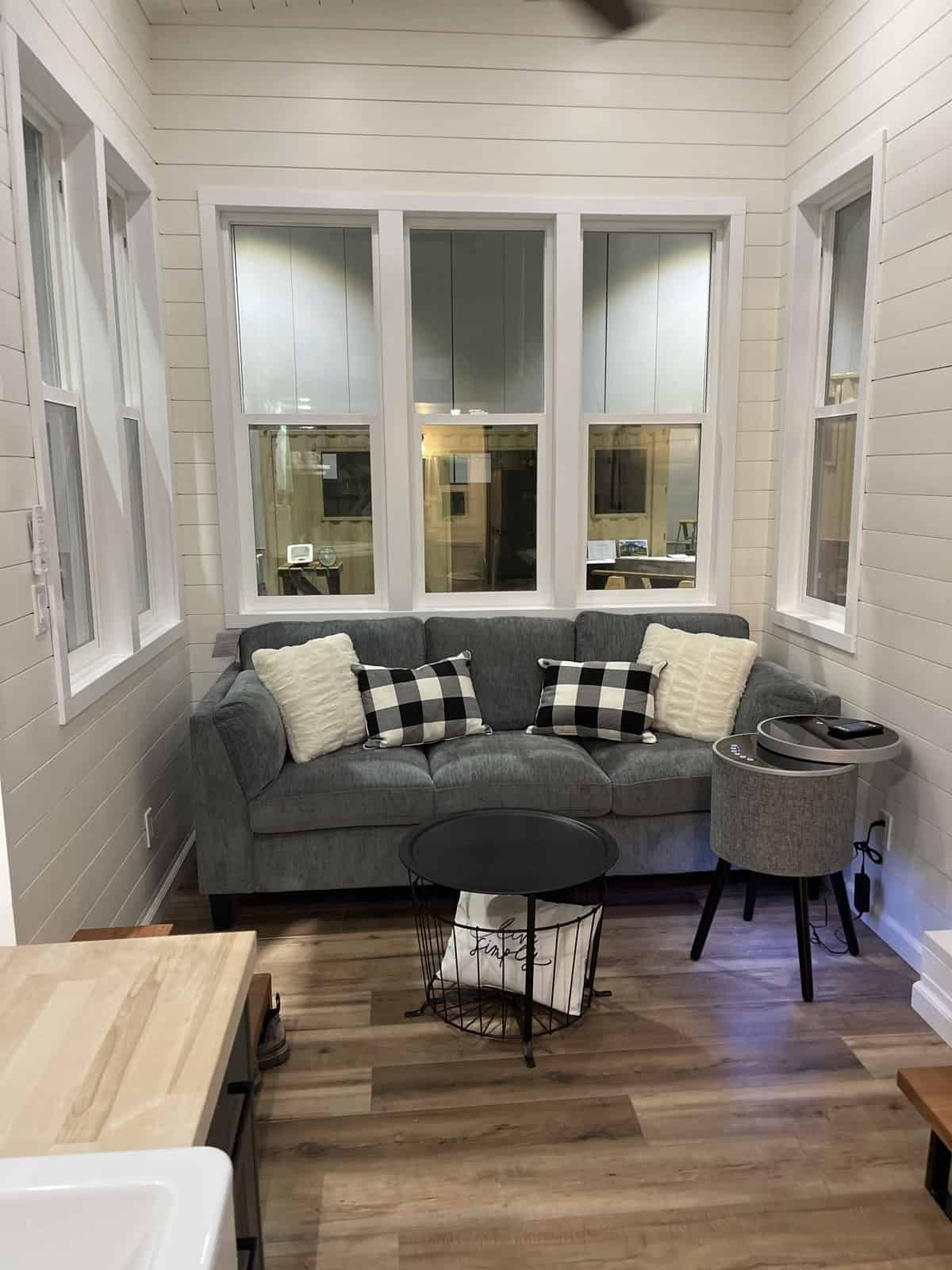 Gray sofa with plaid pillows under a triple window, white shiplap-style walls, and wood-look plank floor