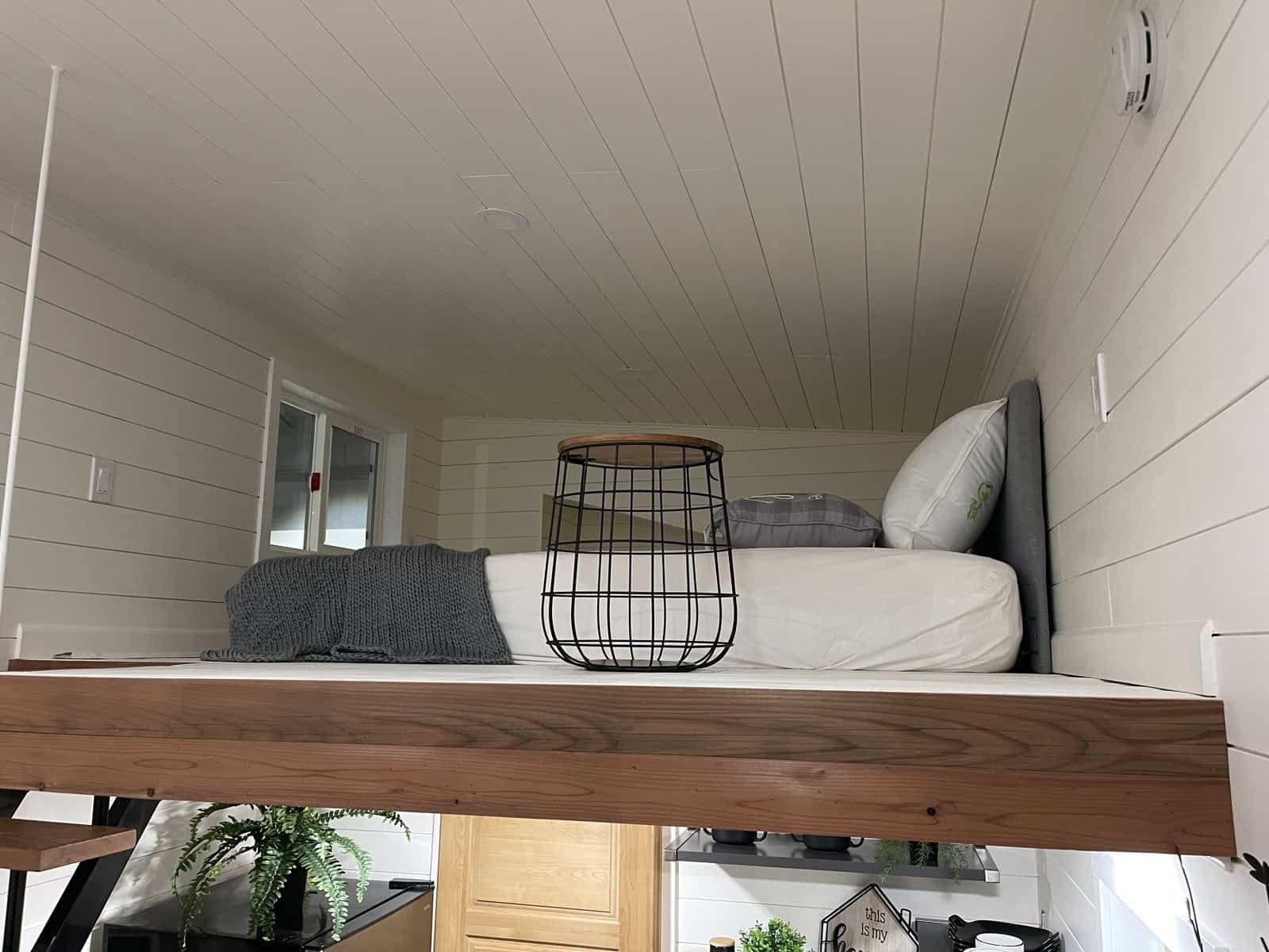 Edge of a sleeping loft with a bed and side table, seen from below, under a white tongue-and-groove ceiling