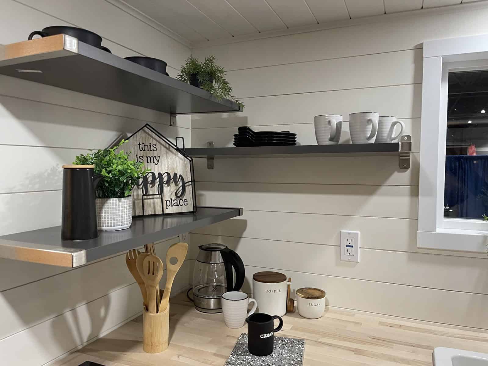 Gray open shelves with mugs and dishes on white shiplap-style walls above a butcher block counter