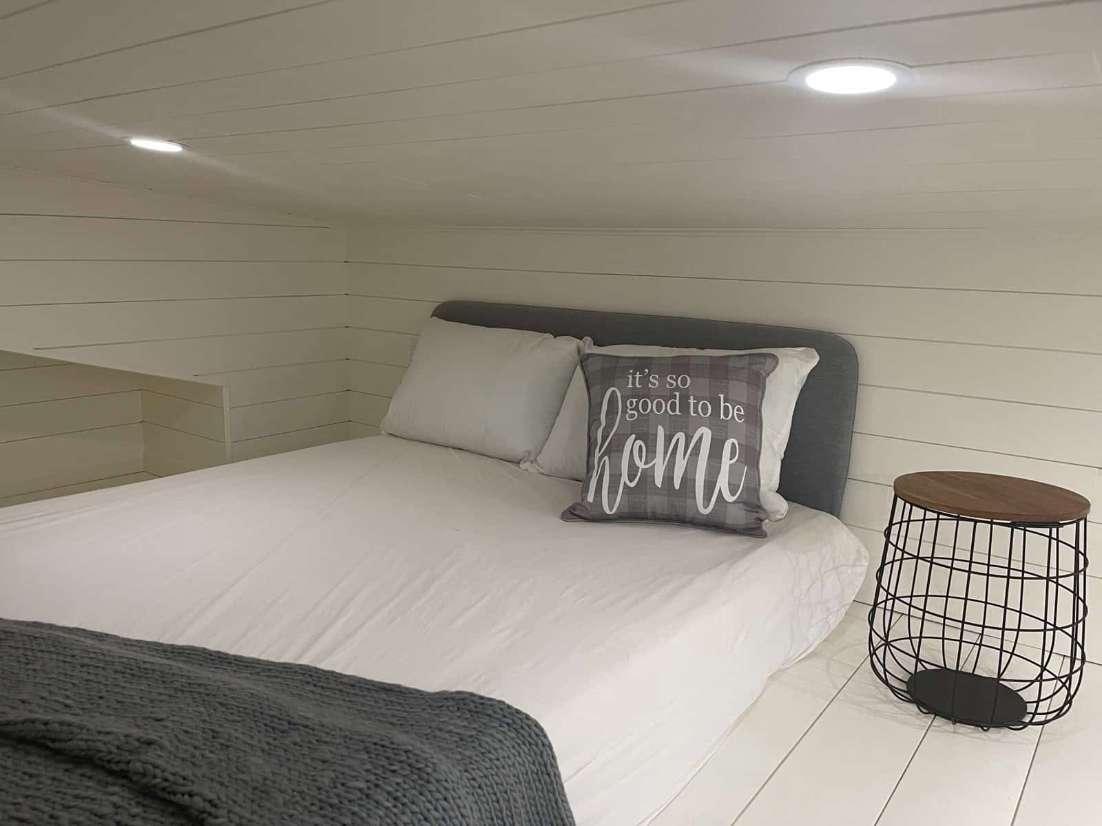 Queen bed with a gray upholstered headboard in a loft under a low white plank ceiling, with a wire side table