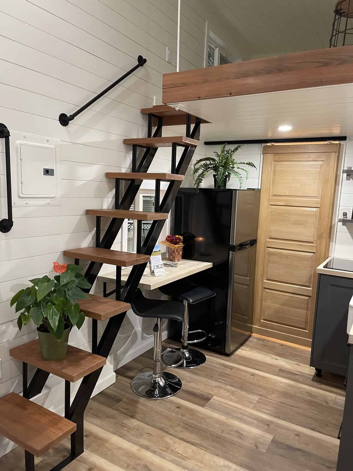 Open-riser wood and black steel stairs beside a small bar counter with two stools, a stainless refrigerator, and a wood barn door