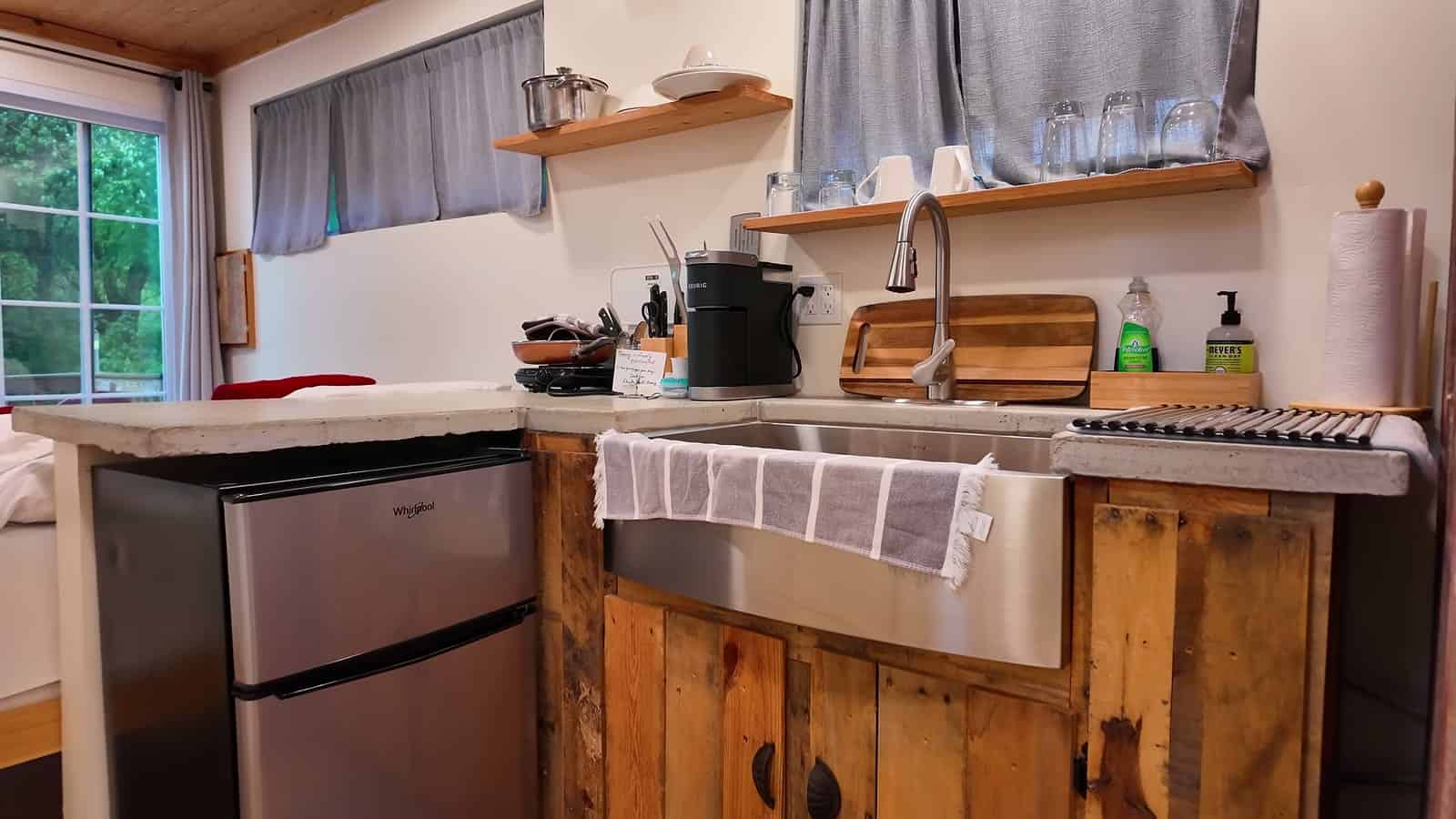 Stainless farmhouse sink above reclaimed wood cabinets and a mini fridge in a container cabin