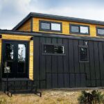 Black tiny house with yellow accents and large windows in a natural setting.