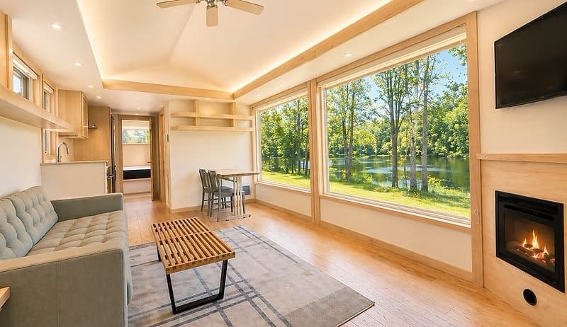 Living room with a gray sofa, electric fireplace, Smart TV, and a wall of glass framing the wooded lake at the Shoreline Lot 17