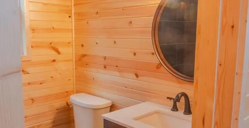Bathroom with knotty pine walls, a toilet, a white vanity with bronze faucet, and a round mirror