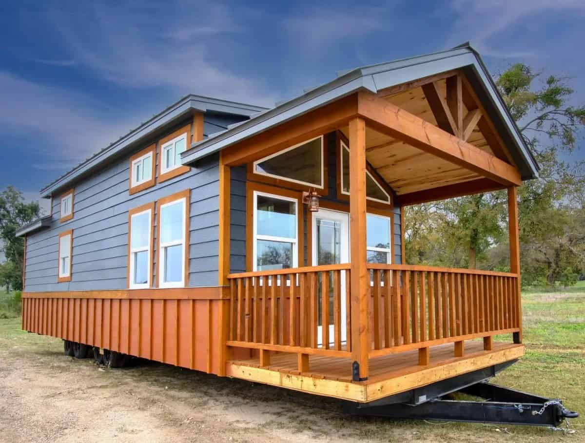 Blue and orange park model with a gabled wood porch and railings, sitting on its trailer