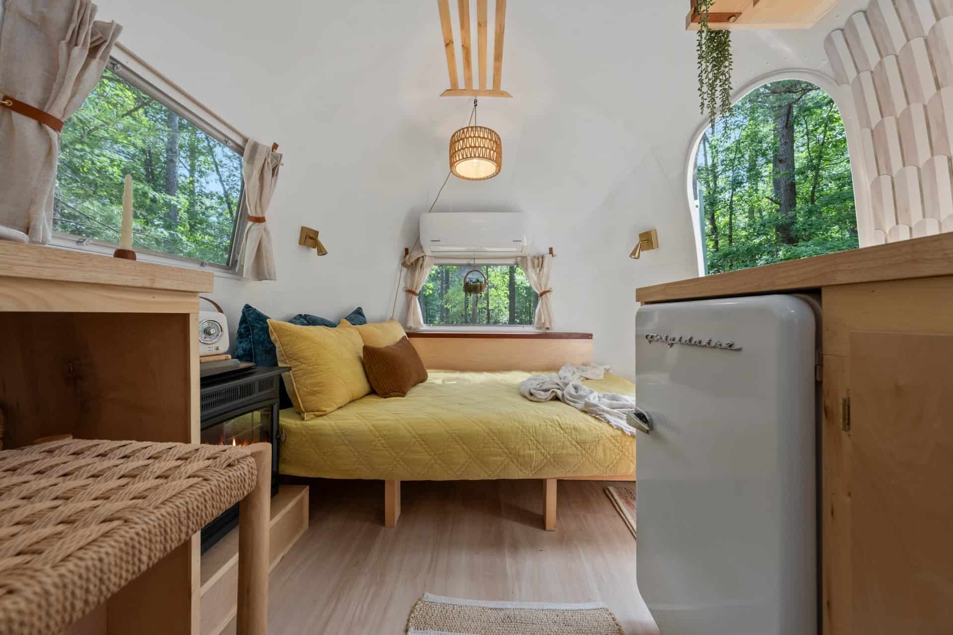 Custom couch in Sol made up as a double bed with a yellow quilt, teal pillows, mini-split above the window, and the kitchen fridge to the right