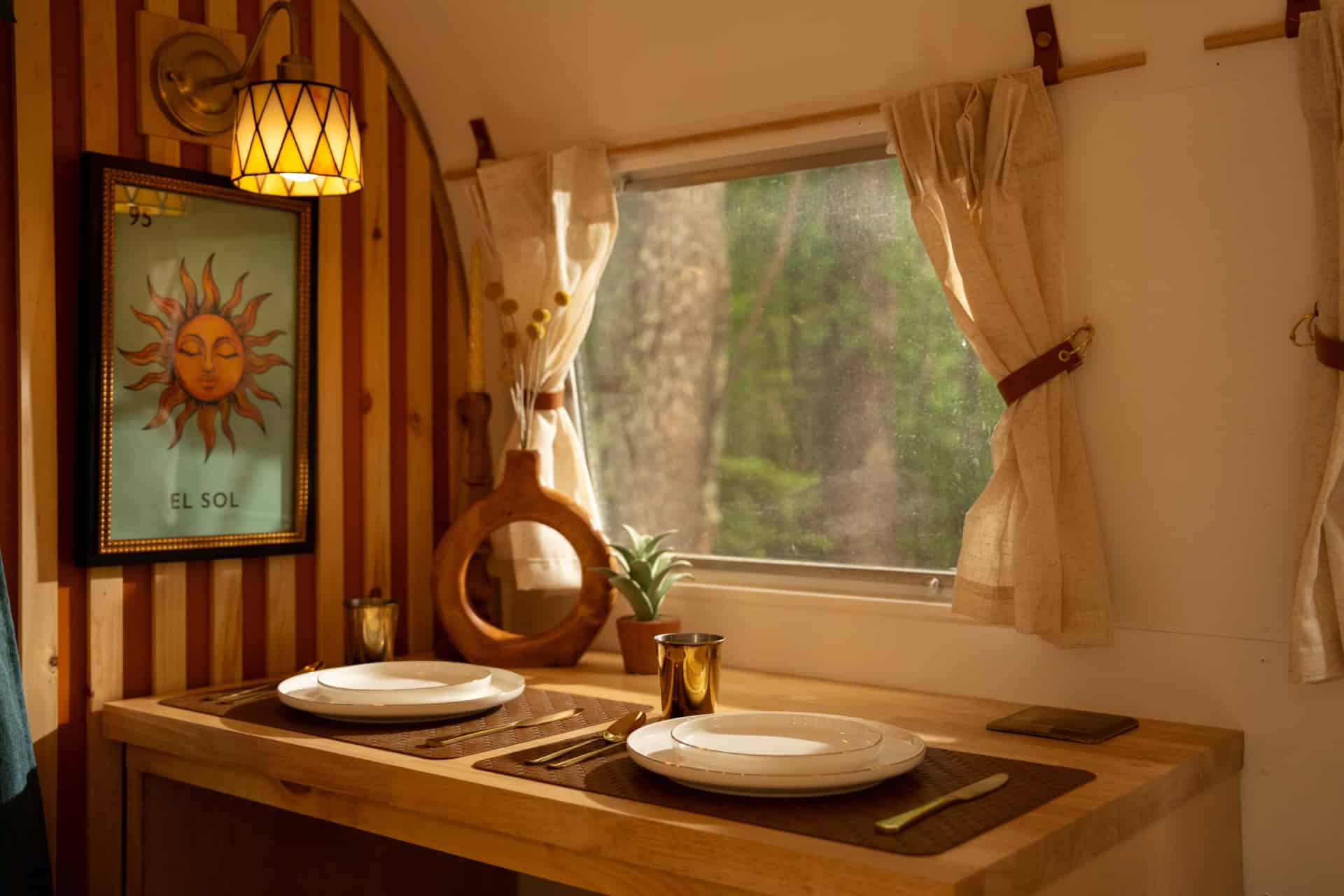 Light-wood dining bar in Sol set for two under a window, with El Sol sun art and a gold sconce on the wood-and-orange slat wall