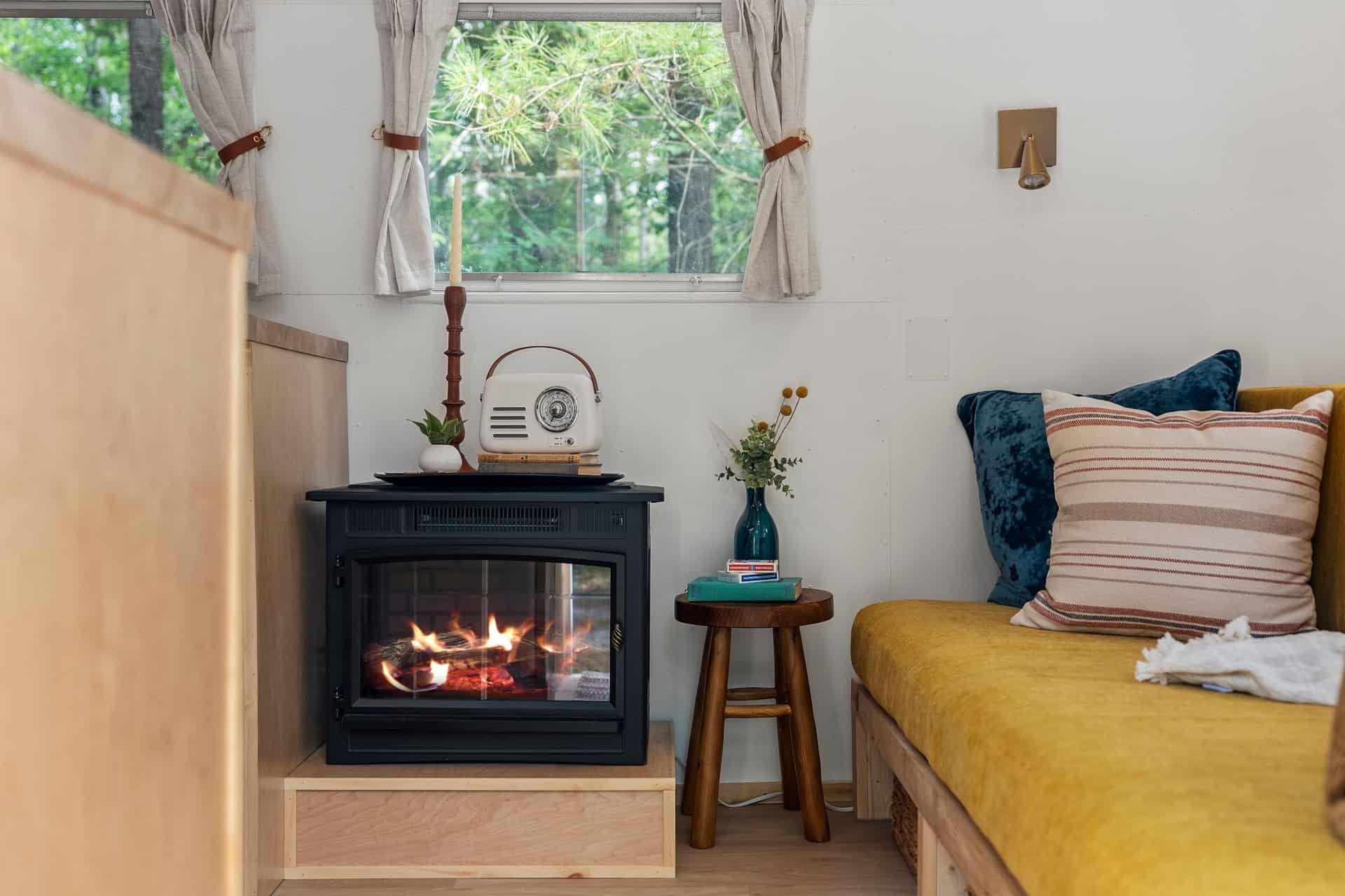 Black electric fireplace glowing beside the mustard custom couch in Sol, with a vintage-style radio on top and woods in the window