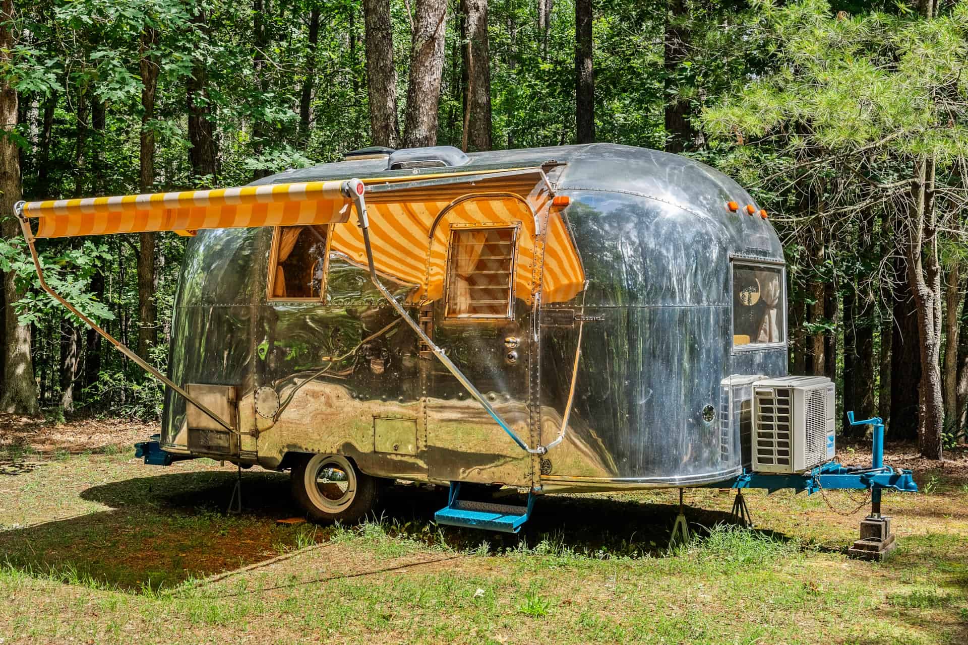 1965 Airstream Caravel Sol with a yellow-and-white striped awning extended, teal hitch, and a white mini-split condenser on the tongue