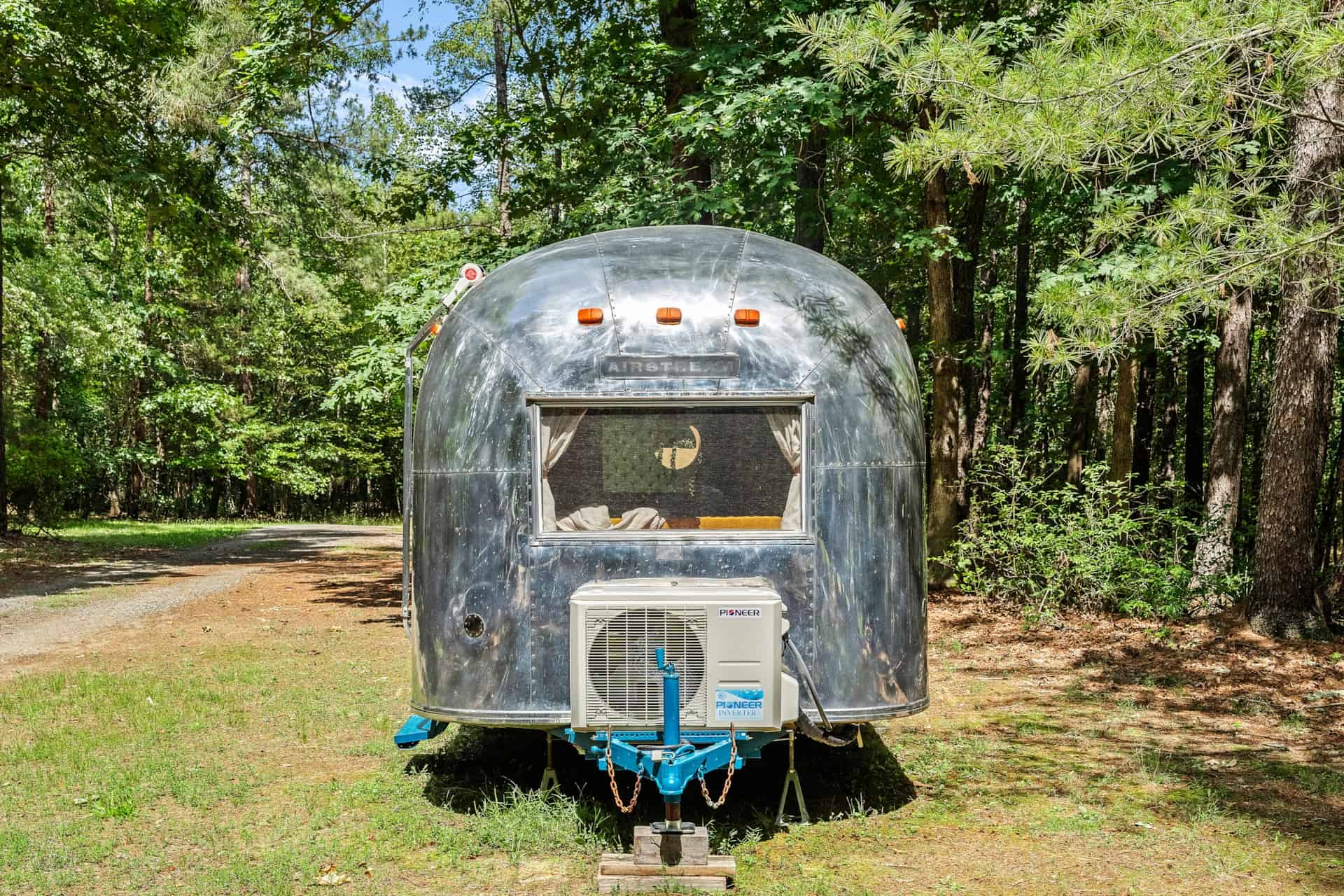Front of the 1965 Airstream Caravel with the AIRSTREAM badge, a Pioneer mini-split condenser on the teal-painted tongue, and woods behind