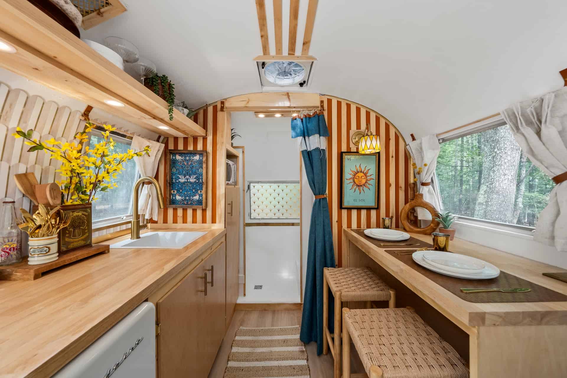 Galley kitchen with butcher-block counter, gold faucet, and wood-and-orange slat wall facing a dining bar set for two under a window