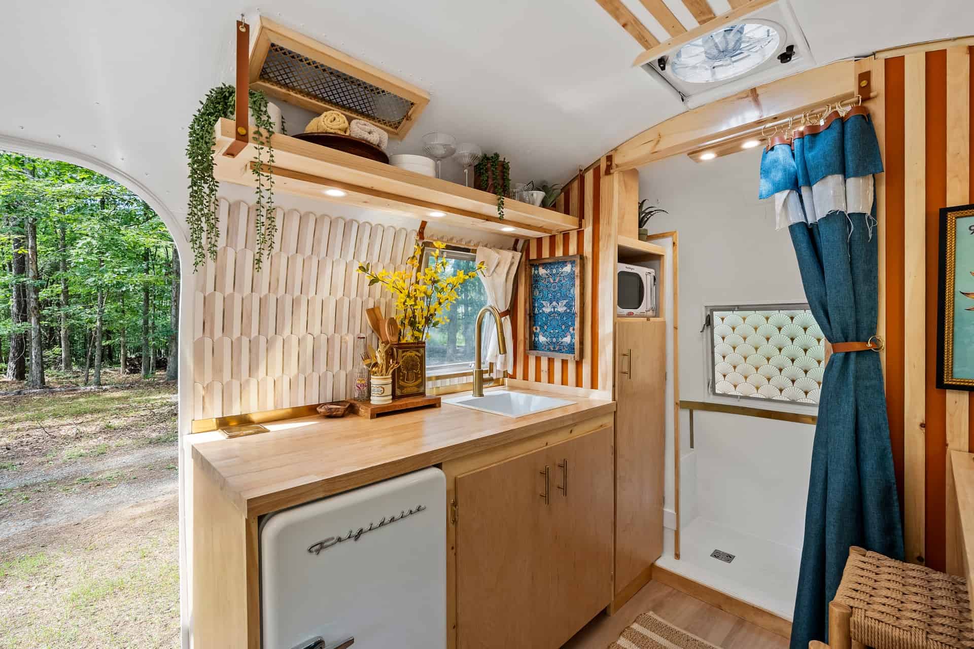 Kitchen of Sol with a white retro fridge under the counter, open wood shelves, a microwave in a pantry cubby, and a tiled shower beyond
