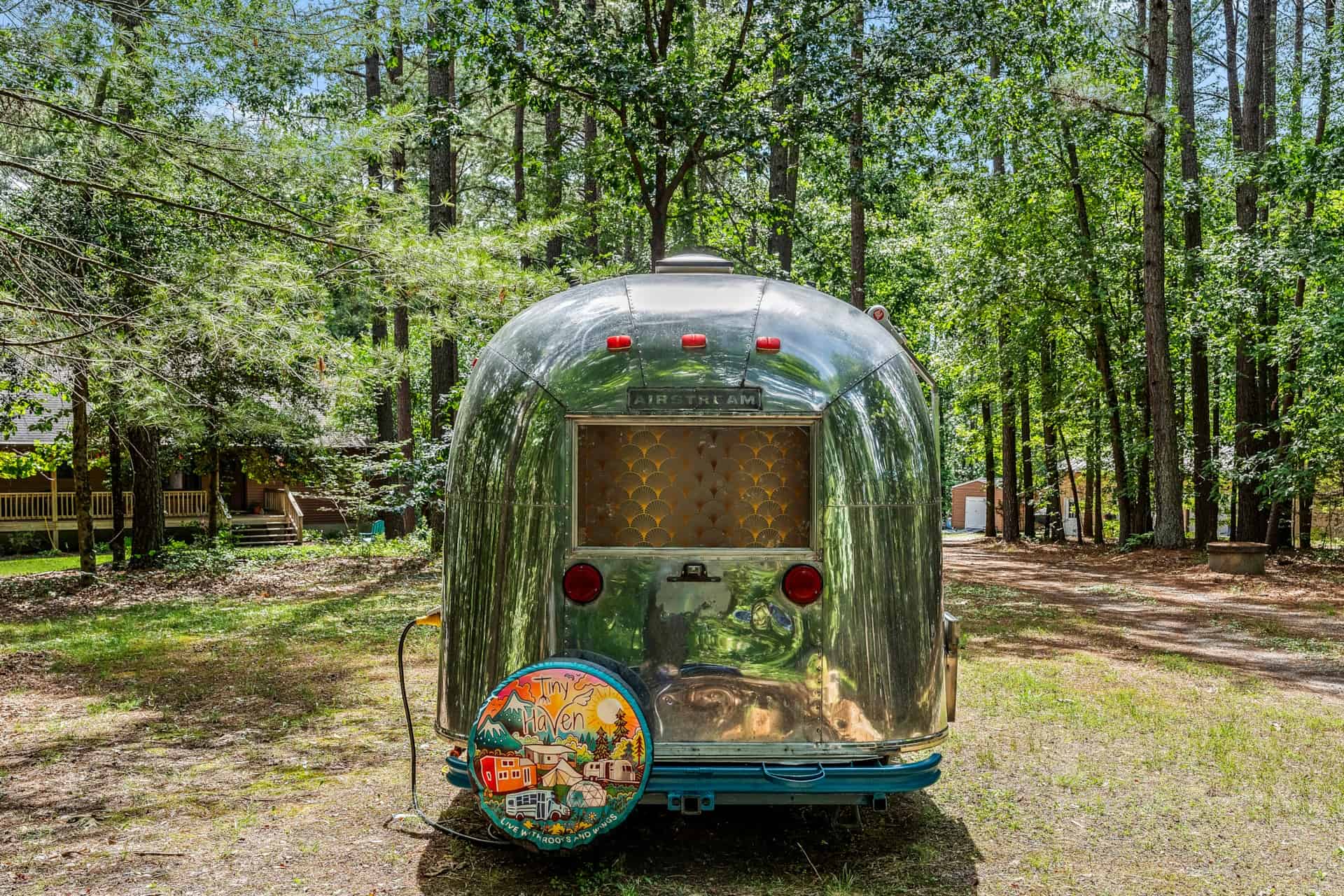 Rear of Sol the 1965 Airstream Caravel with the AIRSTREAM badge, a Tiny Haven spare-tire cover, teal bumper, and a 50-amp cord plugged in