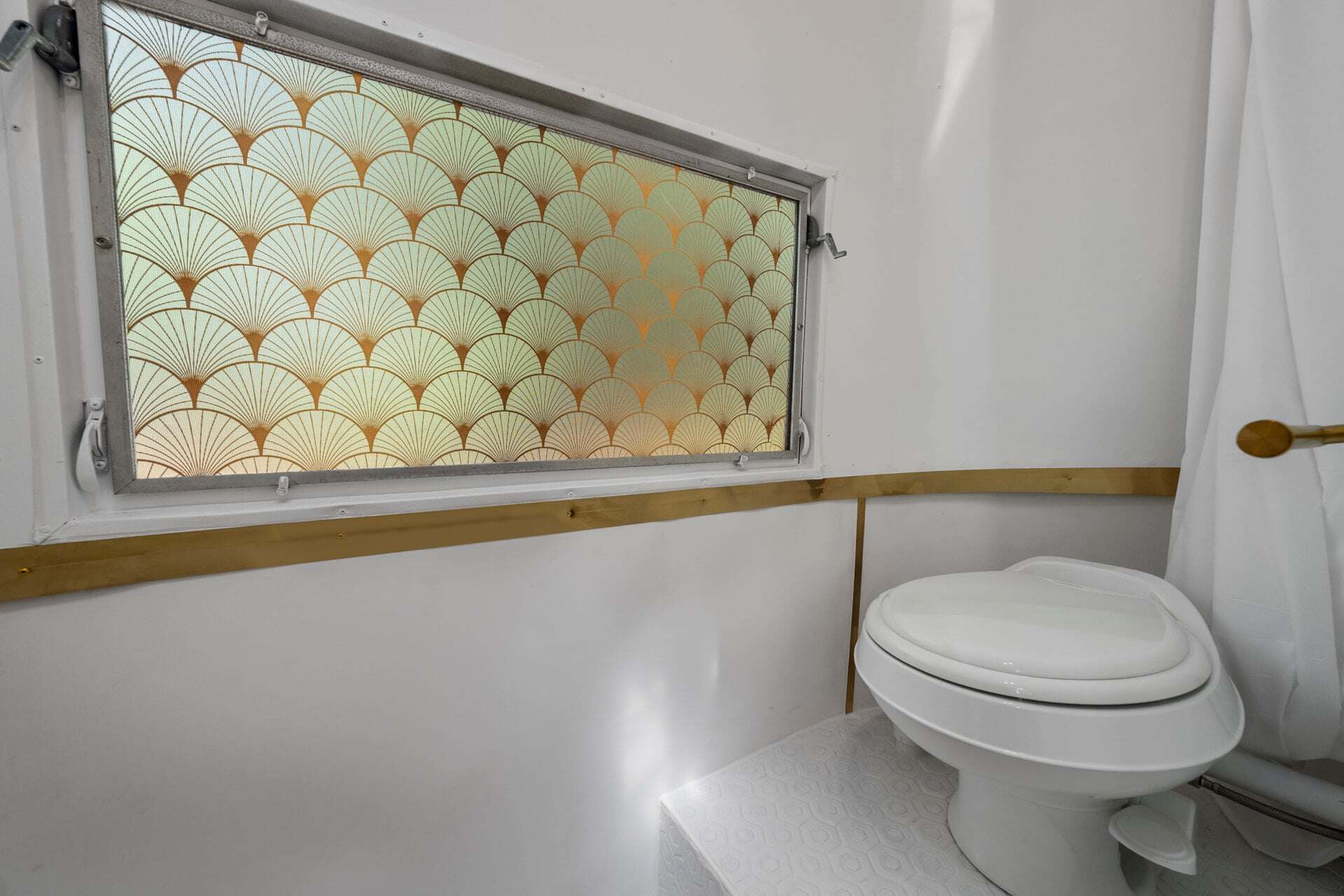 White RV toilet in Sol’s tiled bathroom under a window with gold Art Deco fan privacy film