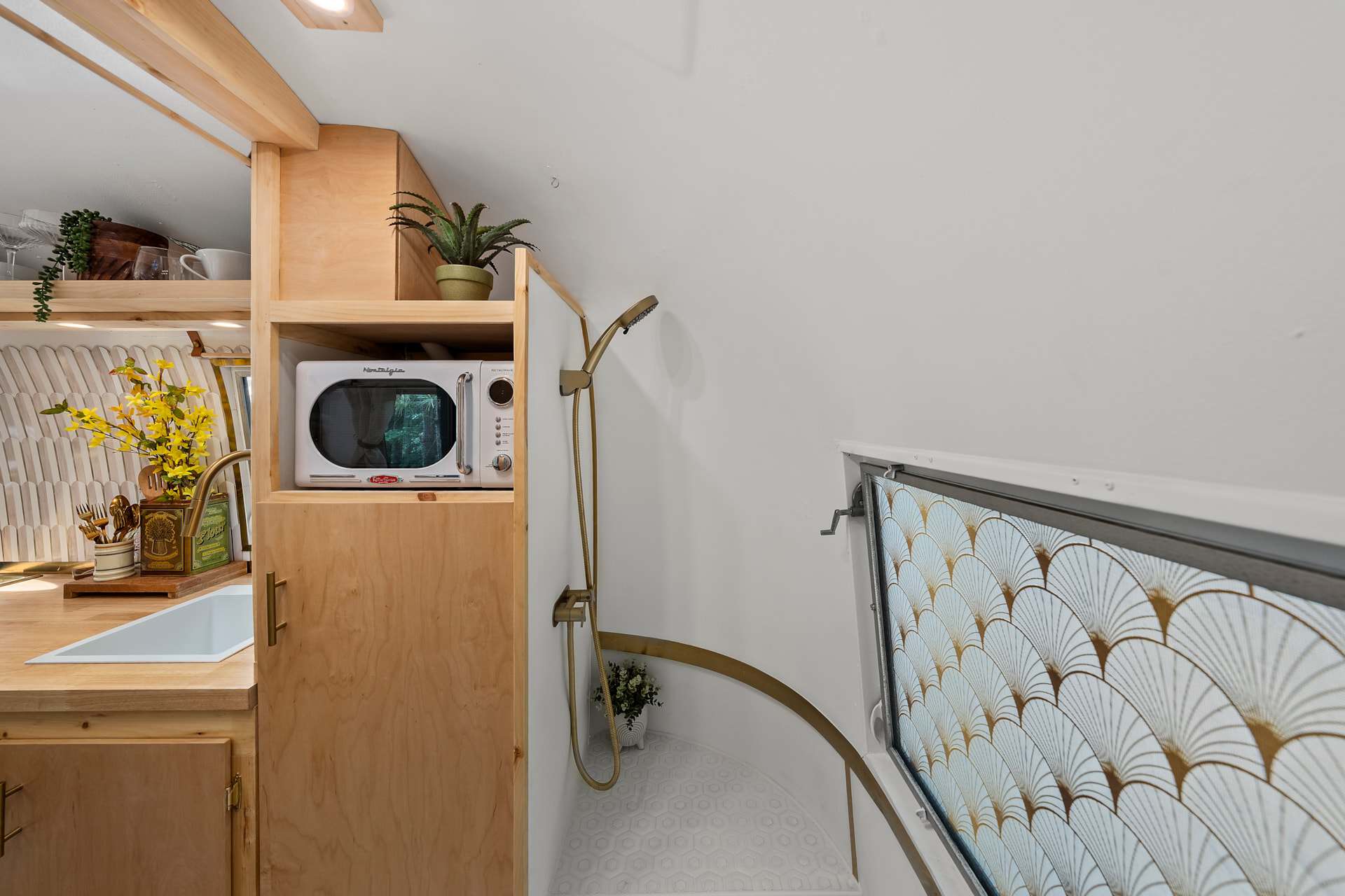 Tiled shower in Sol with a gold shower head and handheld, white hex floor tile, and a microwave cubby in the wood pantry beside it
