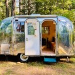 Polished 1965 Airstream Caravel Sol parked in the woods with the door open, a teal hitch and step, and a glimpse of the mustard couch inside