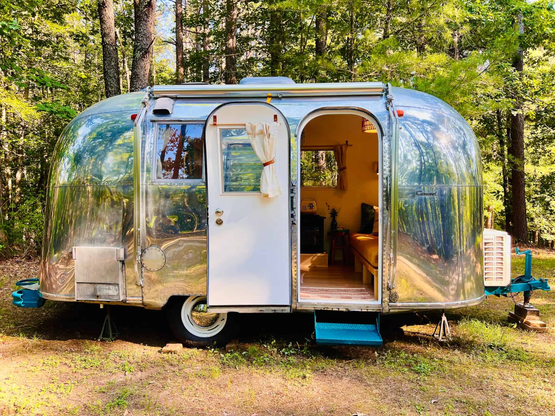 Polished 1965 Airstream Caravel Sol parked in the woods with the door open, a teal hitch and step, and a glimpse of the mustard couch inside