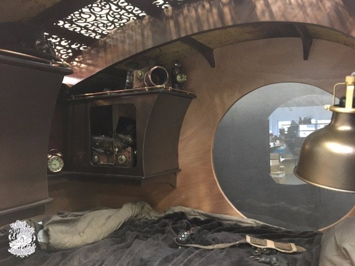 Interior cabin detail with a wall-mounted cabinet, steampunk gauges, brass lamp, and filigree skylight