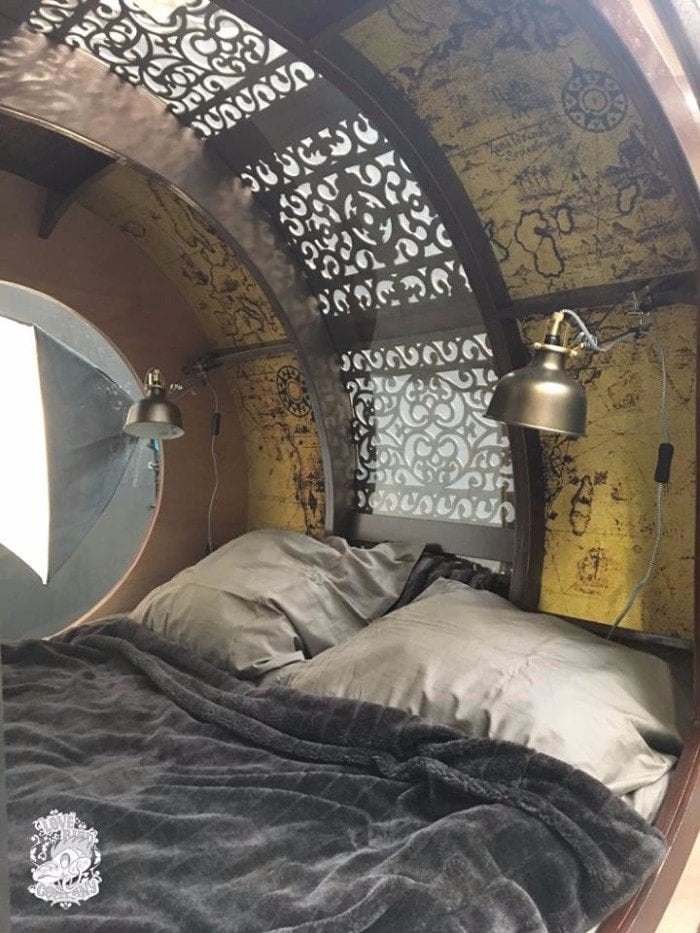 Sleeping cabin of the steampunk teardrop trailer with bedding, two brass lamps, a porthole, and world-map walls