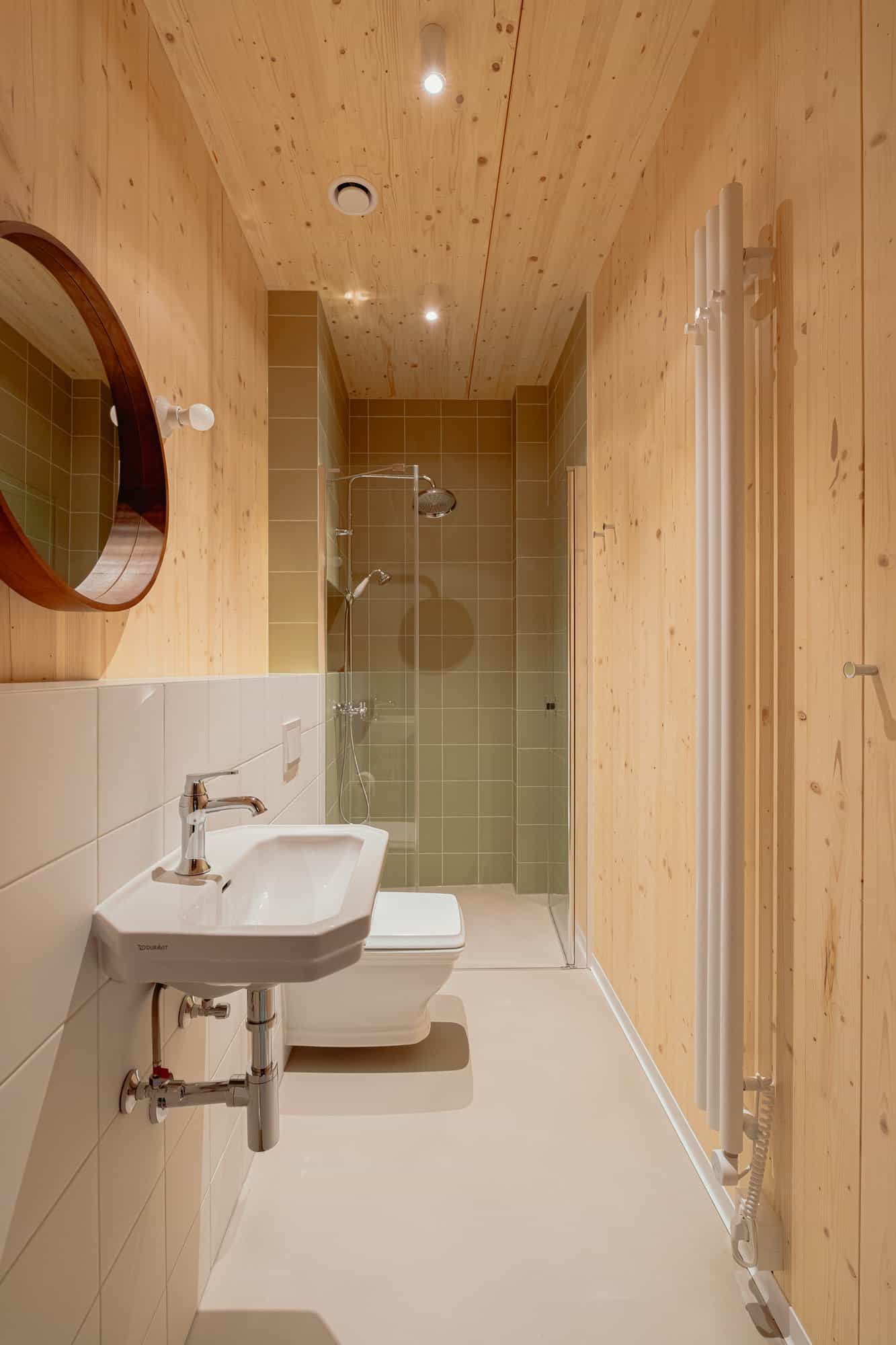 Narrow bathroom with a Duravit sink, toilet, sage-green glass shower, CLT walls, and a white radiator