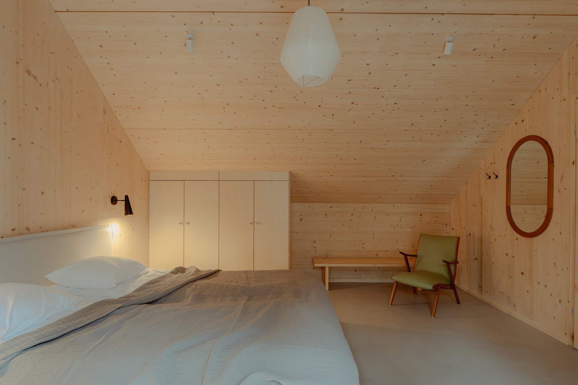 Bedroom with a grey duvet, white built-in cabinets, olive chair, and oval mirror under a sloped CLT ceiling