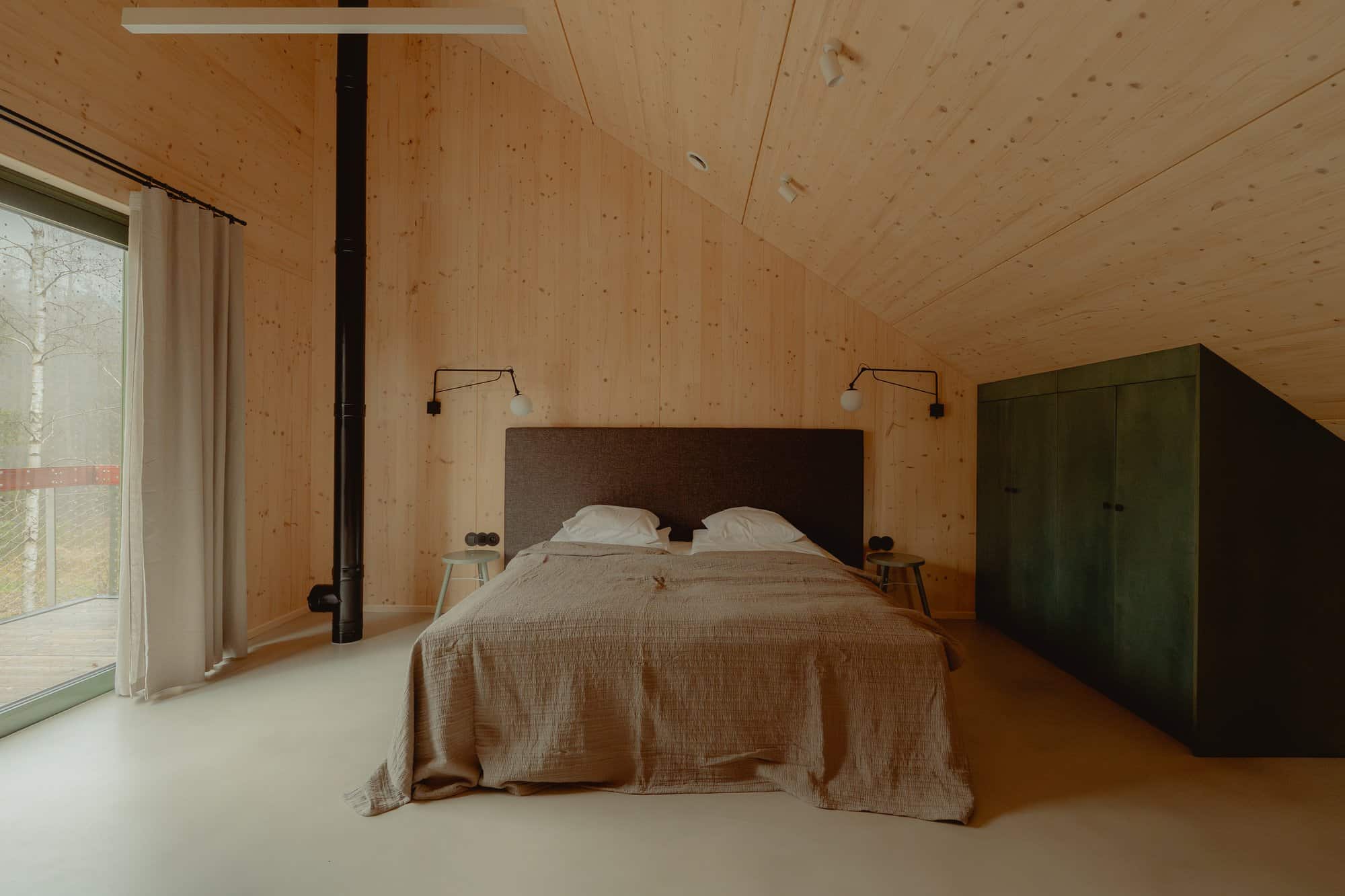 Bedroom under a sloped CLT ceiling with a grey-beige duvet, olive built-in wardrobe, and a forest window