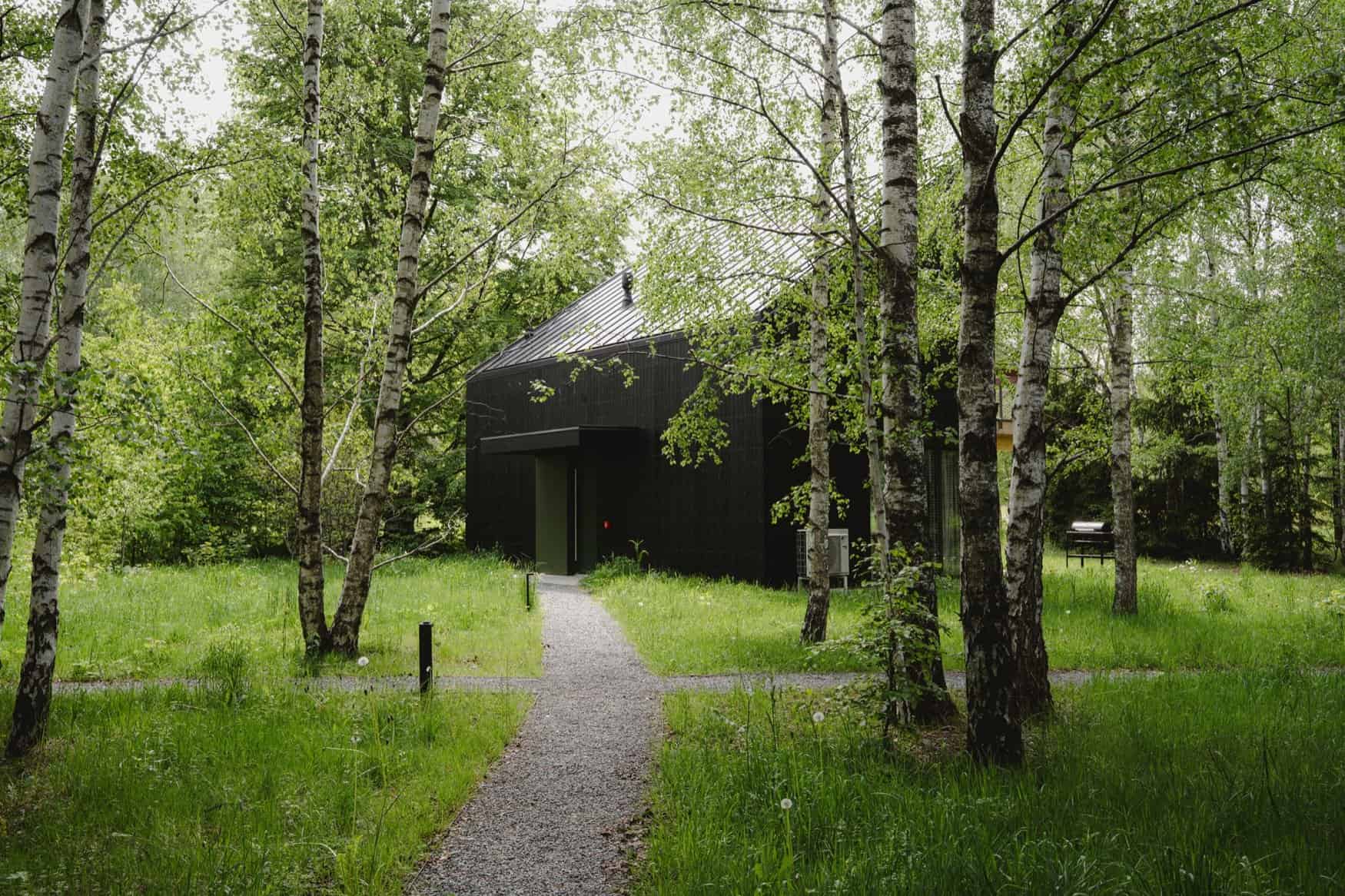 Black cabin at a gravel crossroads with an olive-green recessed entrance, path lights, and a grill in the grass