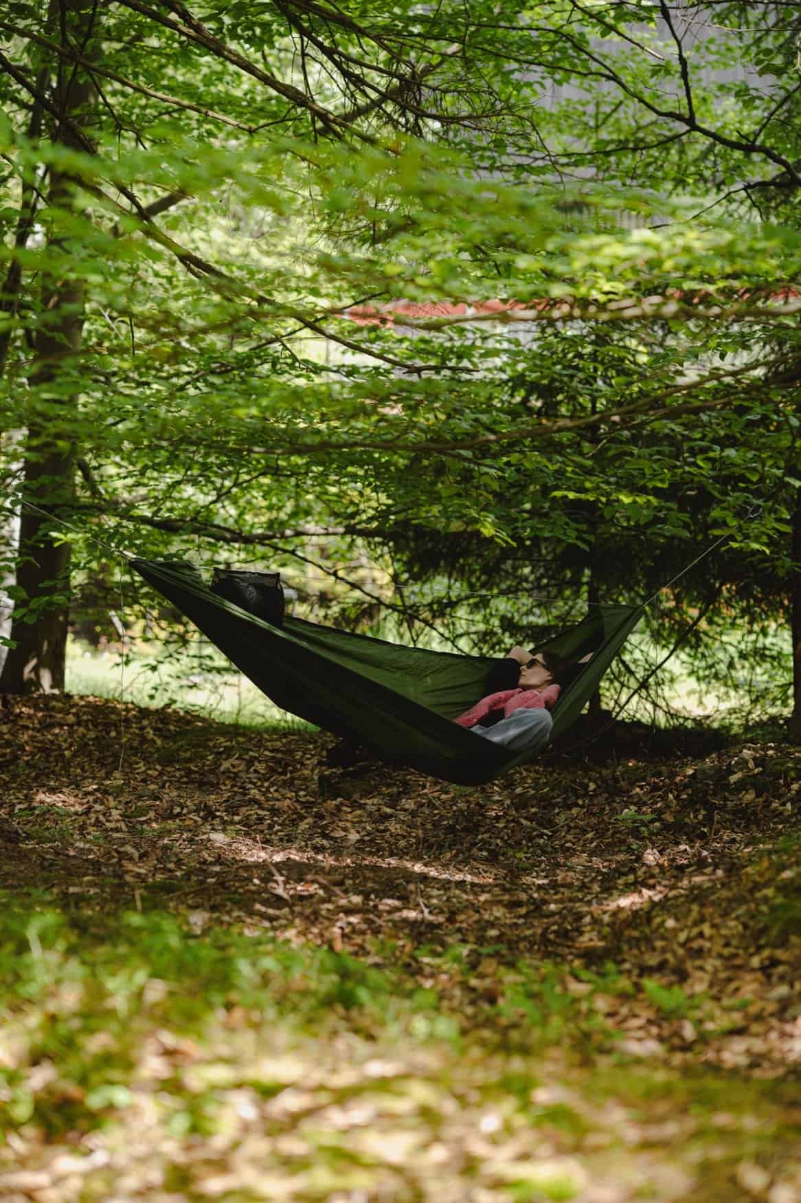 Person in a dark green hammock with sunglasses, a pink garment, and a mesh bag, leaf litter below
