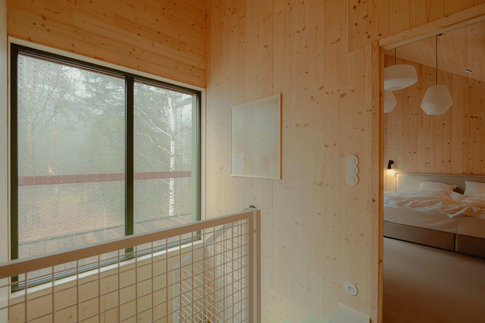 Upstairs landing with a mesh stair rail, birch through a large window, and a doorway into a bedroom with a reading lamp