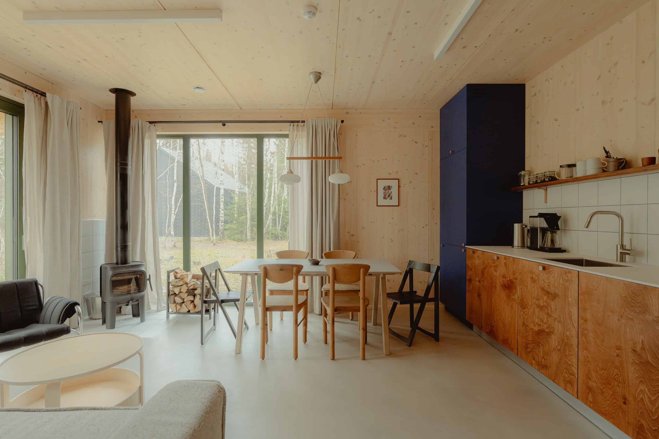 Ground-floor living room of a Studzienno 27 cabin with CLT walls, open kitchen, dining table, and fireplace by the forest glass
