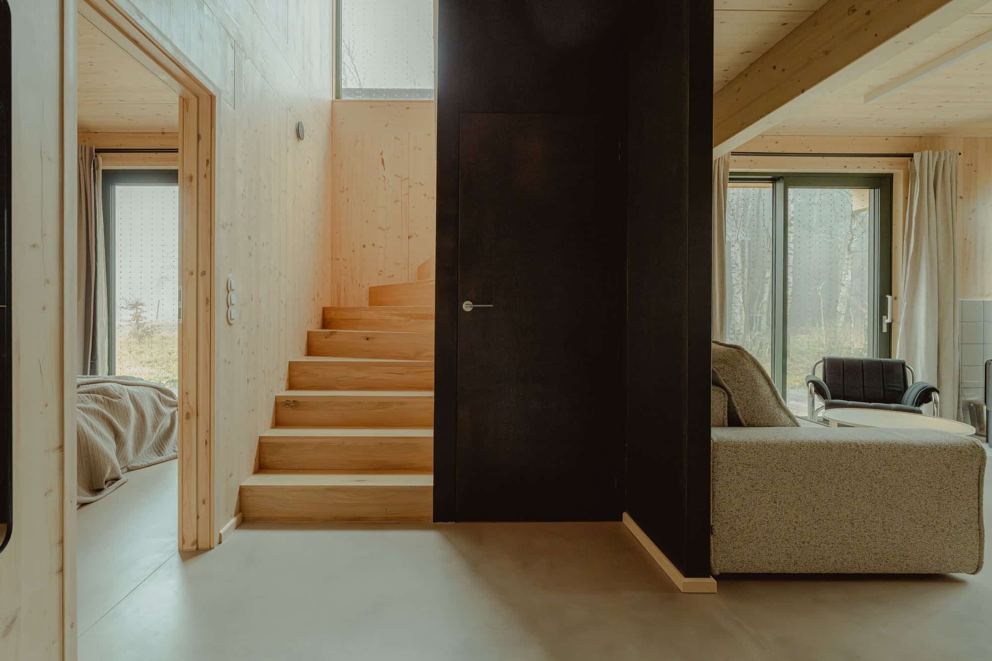 CLT stair, a doorway into a bedroom, and the living room with a sofa and forest glass beyond a black partition