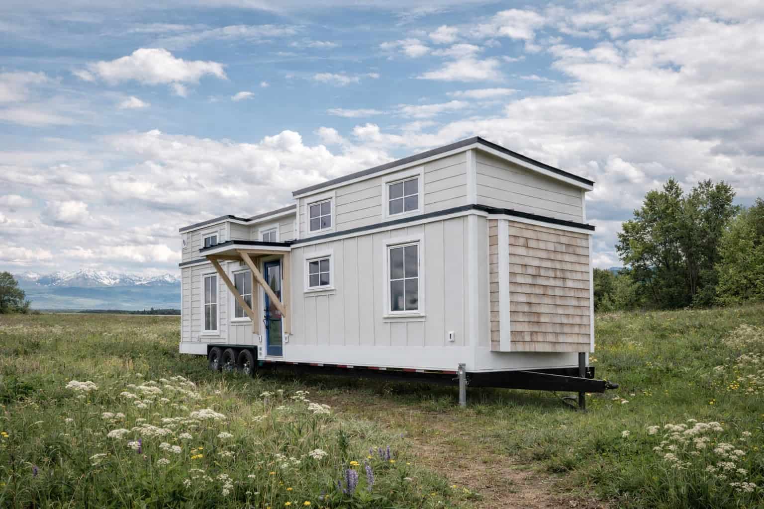 Summit Boho tiny house exterior: white board-and-batten and lap siding, cedar shingle end wall, blue front door under a wood-bracket awning, two loft dormers, triple-axle trailer in a wildflower meadow with mountains behind