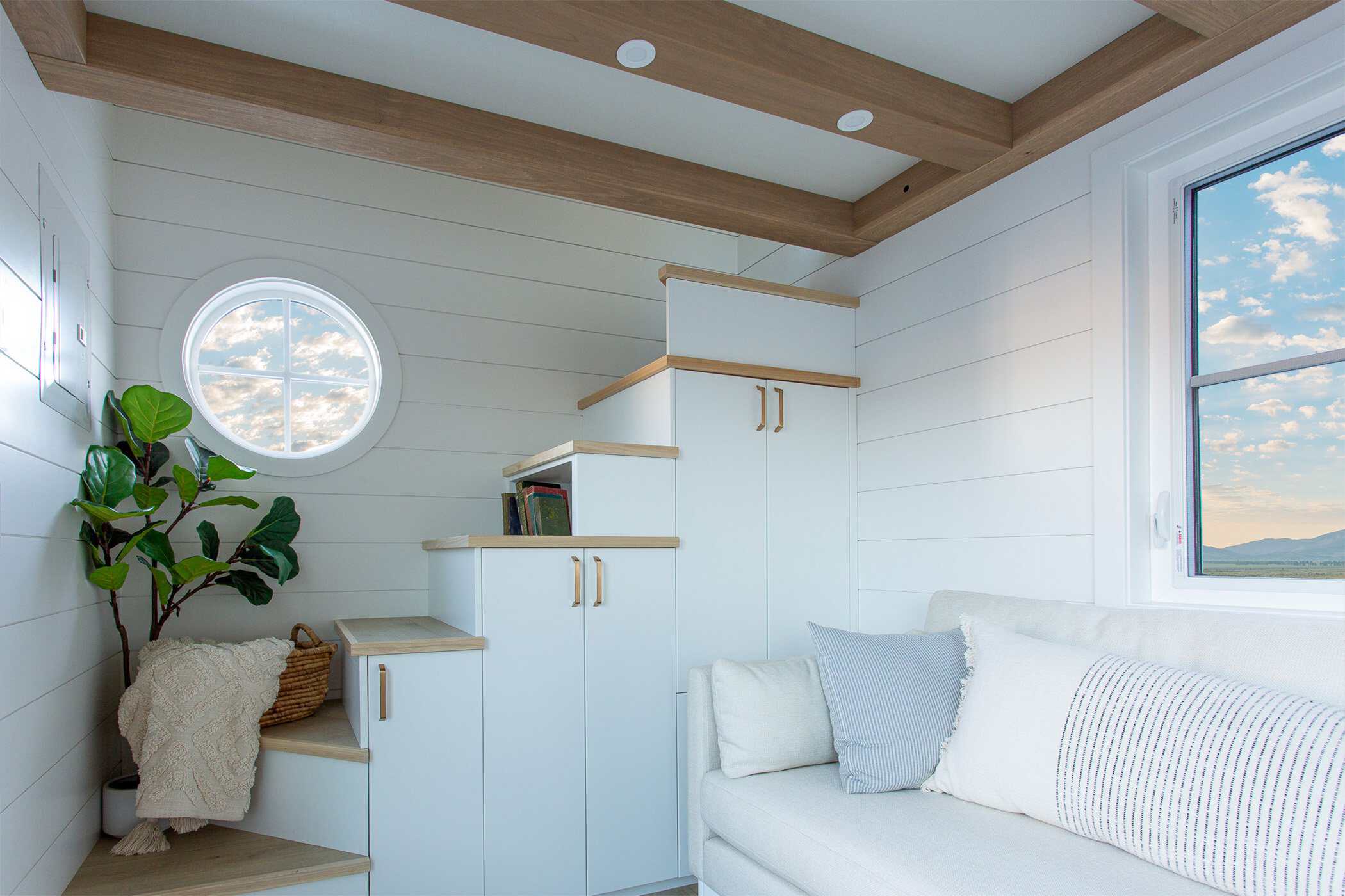 Summit Boho storage stairs with white cabinet fronts under wood treads, white sofa, round window, wood ceiling beams