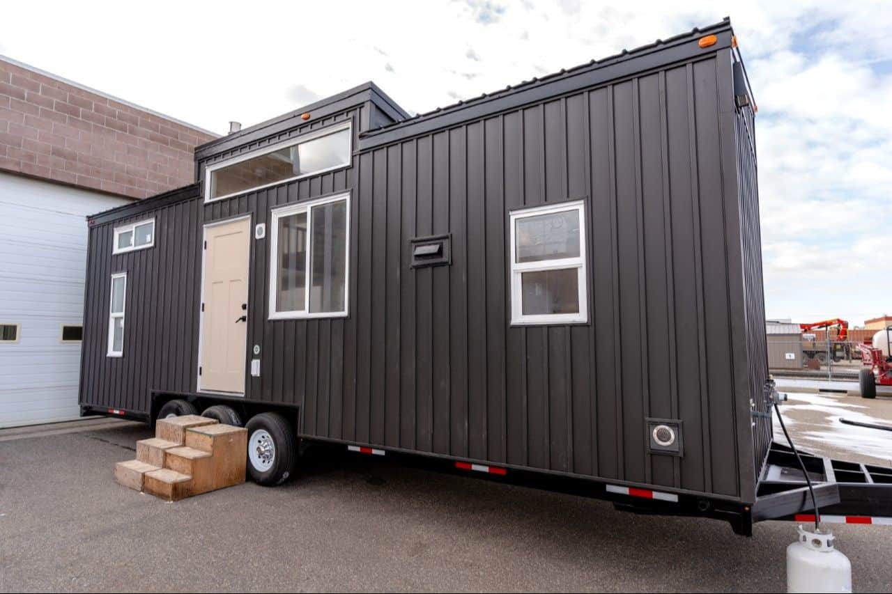 2026 Ruby exterior: black vertical metal, white windows, cream door, stepped shed roof, triple-axle trailer on a shop lot