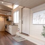 2026 Ruby interior: light shiplap, dark open-riser stair on the left, loft rail over a cream kitchen, craftsman door on the right