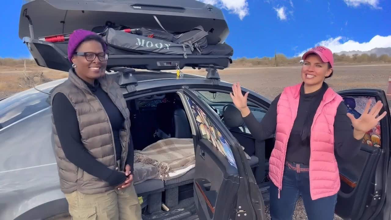 Silver 2005 Nissan Maxima beside desert scrub: Thule roof box open, rear door open showing sleeping platform, Tee in purple beanie and CheapRVliving host in pink vest