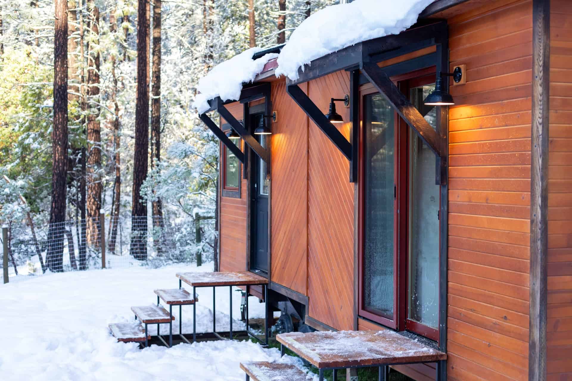 A small wooden tiny house with snow-covered roof in a winter forest setting.