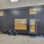 Modern black tiny house on wheels with large window and door, inside visible, in an indoor setting.