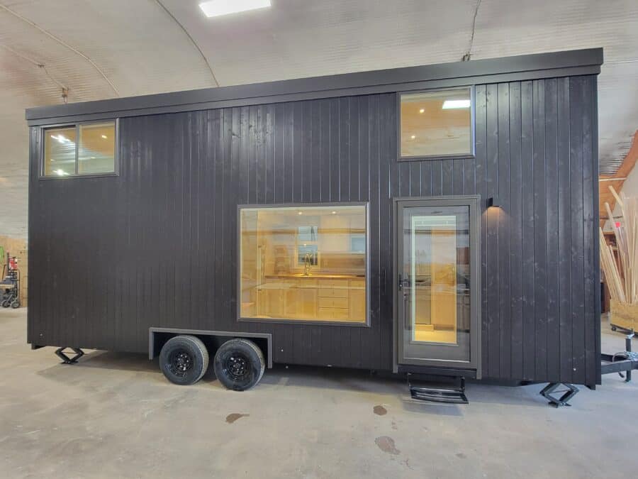 Modern black tiny house on wheels with large window and door, inside visible, in an indoor setting.