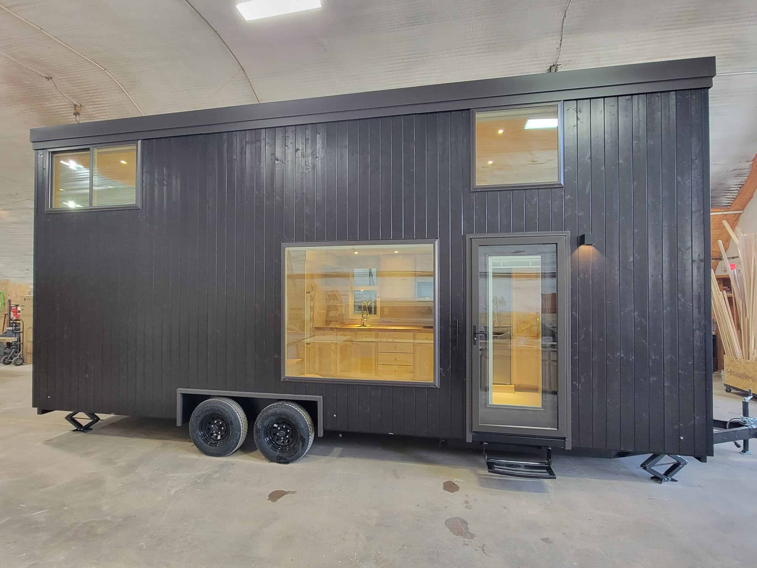Modern black tiny house on wheels with large window and door, inside visible, in an indoor setting.