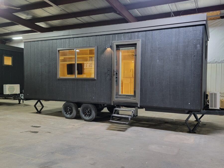 Modern tiny house on wheels with black exterior and large windows inside a warehouse.