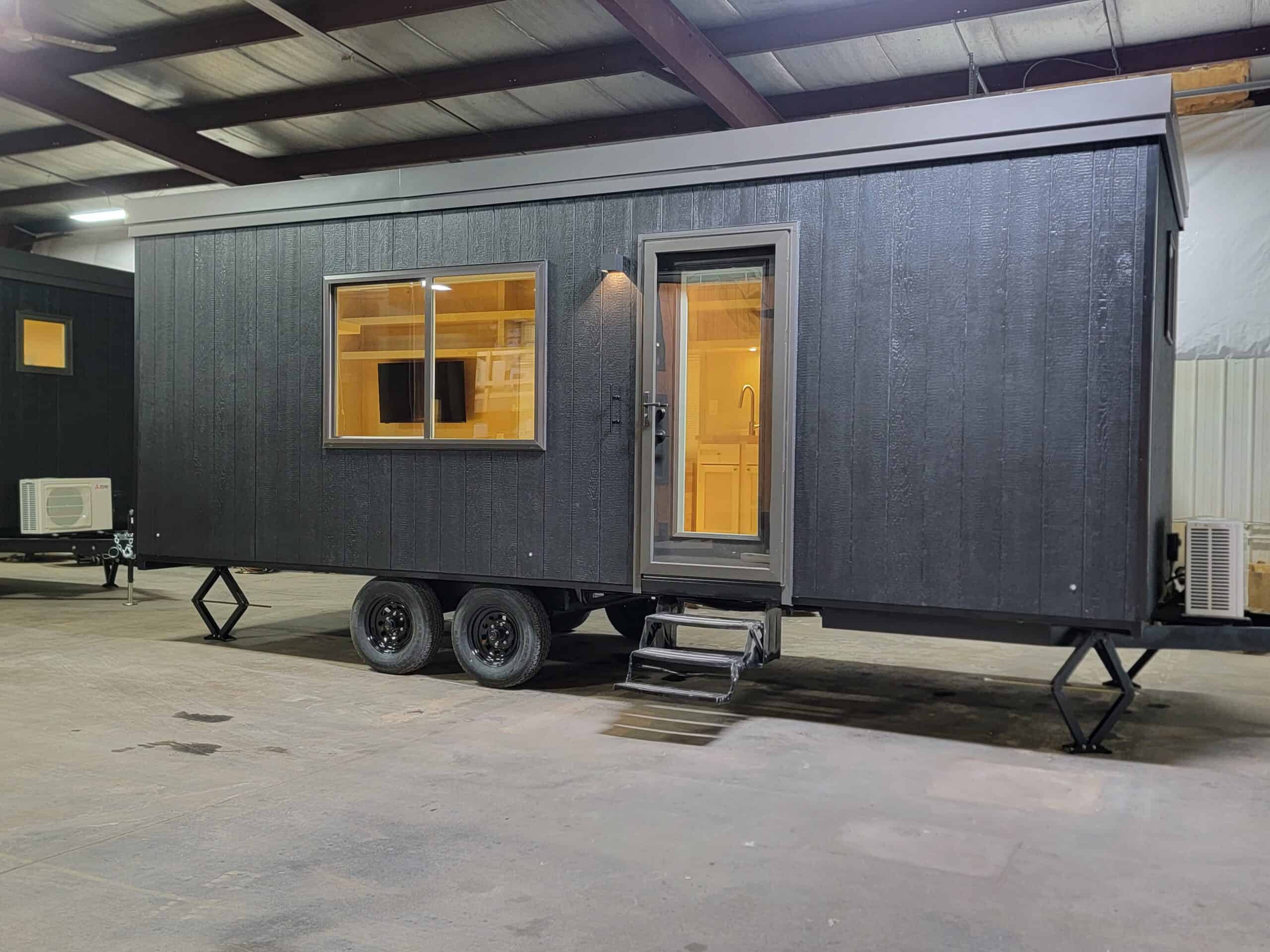 Modern tiny house on wheels with black exterior and large windows inside a warehouse.